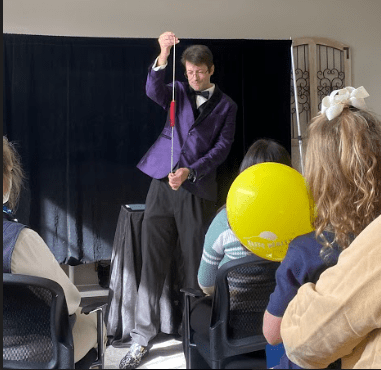 Jordan Allen is holding a wooden heart that moves up and down a string in front of an audience. One audience member has a yellow balloon. 