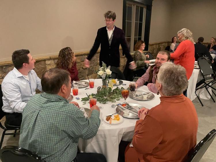 Jordan Allen stands beside a table of eager onlookers at a banquet - awaiting to see what Jordan will do